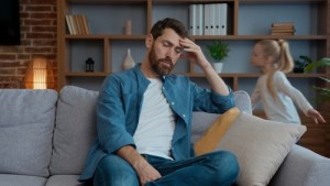 stressed father sitting on couch holding head while child plays behind him