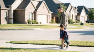 father and young child with backpack walking along suburban neighborhood sidewalk